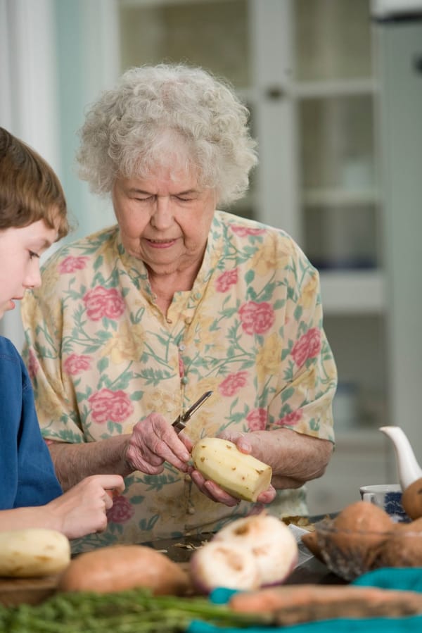 Elderly woman peeling potatoes with young boy looking on.